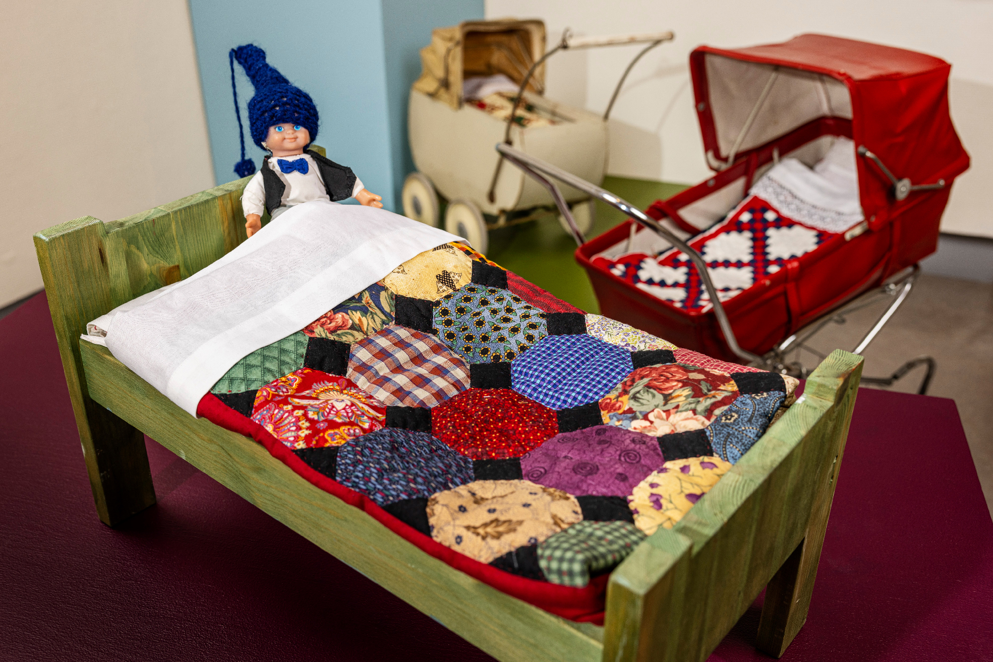 Dockan Nils Holgersson i en docksäng med ett lapptäcke i dockformat. I bakgrunden två äldre dockvagnar med lapptäcken.